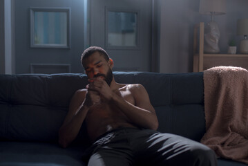 African American man smokes cigarette sitting shirtless on sofa. Deep emotional distress concept