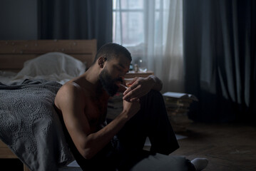 Lonely African American man experiences deep emotional distress sitting on bedroom floor