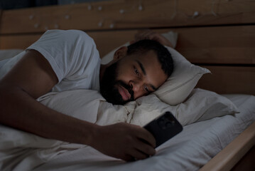 Sleepless black man surfs social media via smartphone lying in bed