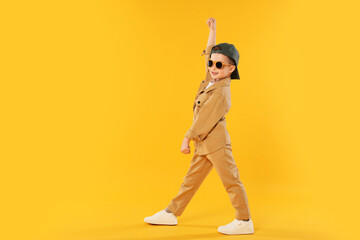 Happy little boy in sunglasses and baseball hat dancing on orange background. Space for text