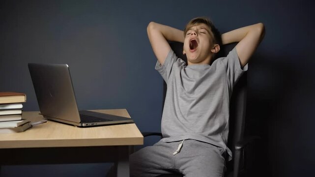 Tiredness and Fatigue: A young student, with hands behind head and eyes closed in exhaustion, taking a break during online class, laptop visible on the wooden desk.