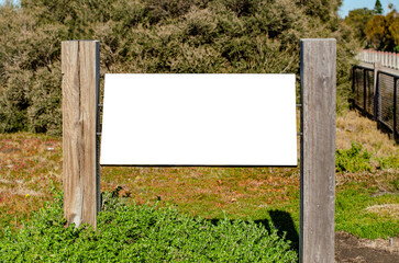 A mockup of a blank white sign mounted on rustic wooden posts in a natural outdoor green space or a...