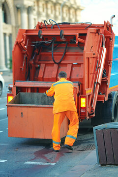 Utility worker in bright orange uniform throws grabage from trash box into garbage truck during waste collection on city street. Sanitation worker loading trash can into utility truck, cleanup routine