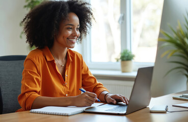Young black woman studies or works at home office using laptop. Female writes in notebook, happy smiles. Remote learning, online course, telecommuting, e-learning, home schooling concept.