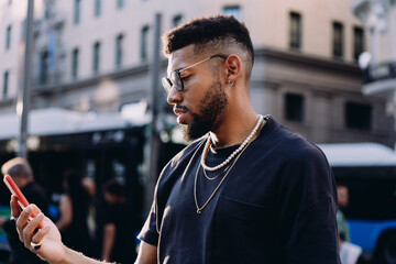 Stylish Black man in his 20s with beard and glasses using smartphone in urban setting, wearing gold and pearl chains, headphones around neck, sunny day with soft natural light