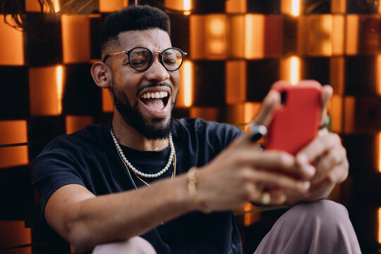 Close-up of African American man laughing while holding smartphone, sitting against orange-lit urban background, possibly reacting to stock market or money transfer success on app