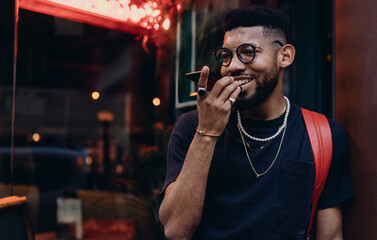 Smiling young Black man using voice command on phone, standing outside vibrant café at night,...