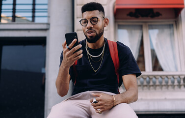 Young African-American man interacting with phone, seated in front of urban facade, stylish and...