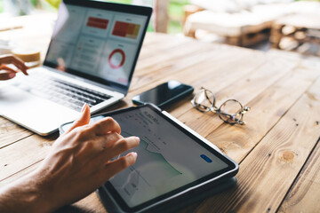 Hands using tablet for real-time financial stats next to open laptop with business charts, glasses...
