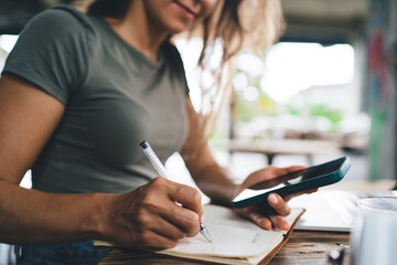 Freelancer writing in notebook while referencing smartphone, smiling and focused in cozy outdoor...