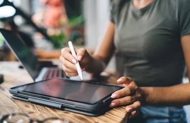 Close-up of young woman using stylus on tablet while working remotely with laptop, managing cryptocurrency or financial analytics in cozy modern environment with natural daylight