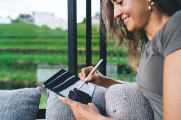Smiling digital nomad woman drawing with stylus on tablet while relaxing on cushion by the window,...