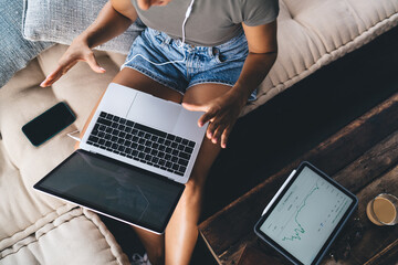 Woman multitasking with laptop on lap, sitting on sofa, using earphones while tablet with trading...
