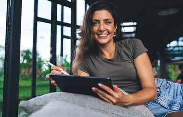 Smiling woman lying on sofa with tablet and stylus, looking at camera, enjoying relaxed freelance...