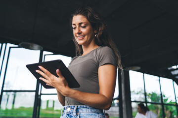 Confident woman standing indoors using digital tablet, smiling while checking or creating content,...