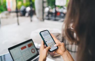 Young freelancer texting on smartphone while managing cryptocurrency and business analytics on laptop, multitasking between messaging, charts, and financial platforms