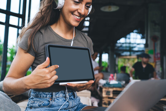 Young woman in denim shorts and headphones smiling as she demonstrates blank tablet screen to laptop during remote video call in modern coworking area with people in background