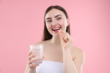 Happy woman with glass of water taking collagen pill on pink background