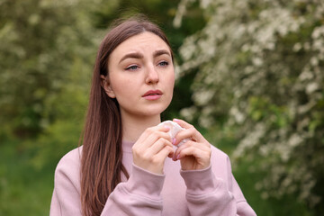 Woman with paper tissue suffering from seasonal pollen allergy near blossoming shrub on spring day