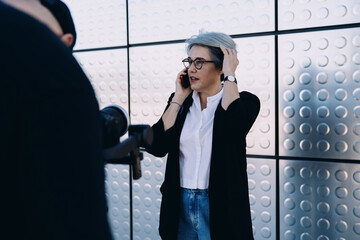 Candid shot of confident mature businesswoman with gray hair adjusting hair while talking on smartphone during outdoor interview or video shoot near modern architecture