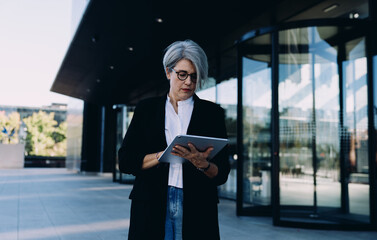 Focused older businesswoman using digital tablet in front of modern office building, managing tasks...