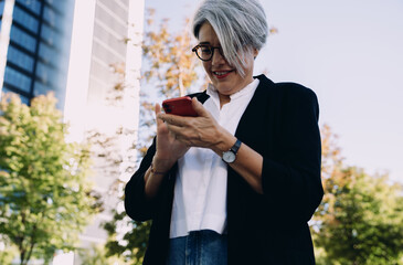 Confident middle-aged trader checks financial data on mobile app while standing outside modern building, representing smart investing, independence and fintech lifestyle