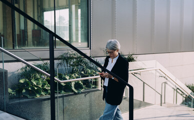 Focused mature female entrepreneur reading smartphone screen while descending office stairs,...