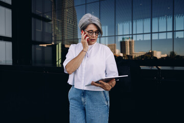 Senior businesswoman with digital tablet talking on smartphone, standing against glass facade,...