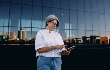 Confident older female freelancer managing tasks on tablet while standing by glass building,...