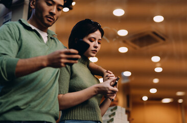 Couple focused on phones while standing indoors with modern lights. Urban routine of travelers...
