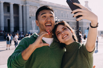 Smiling young interracial couple taking selfies in front of historic architecture on sunny day in Madrid, enjoying romantic vacation and capturing joyful travel memories with smartphone
