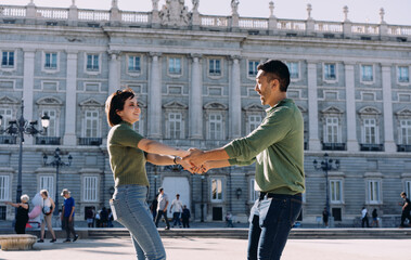 Cheerful couple laughing and holding hands in front of majestic architecture. Playful interaction...