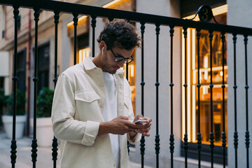 Caucasian confident Man with smart glasses and wireless earphones focused on smartphone. Mobile technology, digital connection, and online engagement concept in illuminated city environment