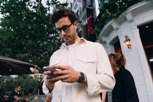 stylish young man in glasses and beige shirt focuses on his smartphone while standing outside chic café. His wireless earbuds and wristwatch reflect a modern, tech-savvy lifestyle in an urban setting