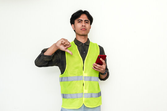 Disappointed Asian construction worker holding smartphone while showing thumbs down gesture, isolated on white background. Perfect for negative reviews or bad service advertisement themes