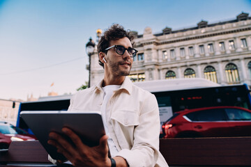 confident male digital nomad sits on city bench, working on his tablet. With wireless earbuds and a...