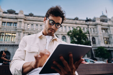 Focused businessman working on tablet in an urban setting. Wearing stylish glasses and wireless...