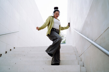 Happy Hispanic woman dances on outdoor stairs wearing headphones, holding a smartphone. Female...