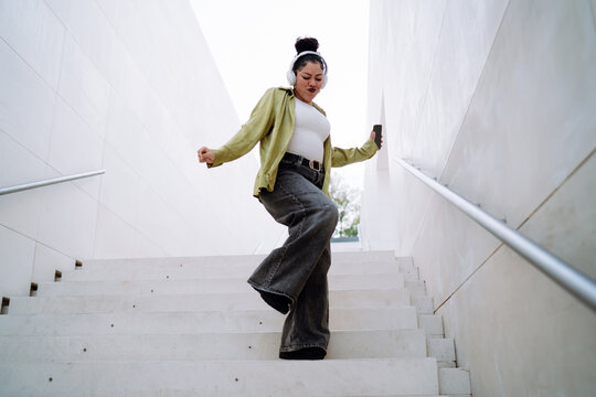 Happy Hispanic woman dances on outdoor stairs wearing headphones, holding a smartphone. Female enjoys music, moving freely with confidence. Digital entertainment and self-expression concept - Powered by Adobe