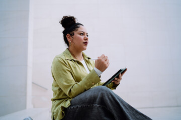 Thoughtful young woman holding stylus and tablet, immersed in creative work. Her focused expression...