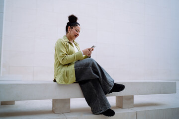 Young woman sits on modern bench, smiling while browsing her smartphone. Female enjoys digital connectivity in minimalistic space, representing online communication, social media, and remote work