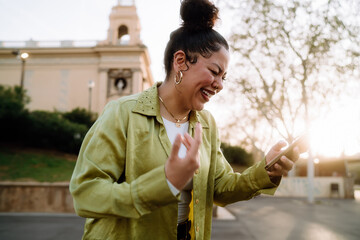 Hispanic woman laughs joyfully while looking at smartphone in outdoor setting. Dressed in green...