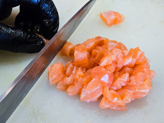 Chef slicing raw salmon into small cubes on a white cutting board, wearing black gloves for hygiene, preparing ingredients for sashimi or poke dishes.