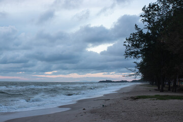 A quiet beach with strong waves under a cloudy sky in the early evening, with pine trees lining the coast and soft light peeking through the clouds