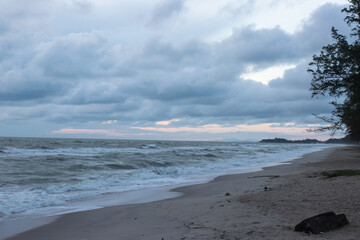A quiet beach with strong waves under a cloudy sky in the early evening, with pine trees lining the coast and soft light peeking through the clouds