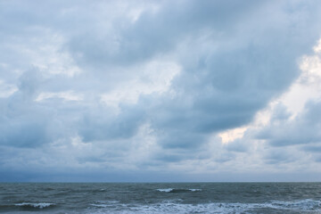 A quiet beach with strong waves under a cloudy sky in the early evening, with pine trees lining the coast and soft light peeking through the clouds