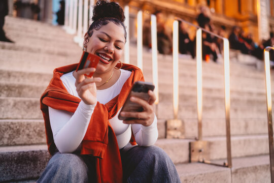 young woman with bright smile enjoys an effortless online shopping experience. Seated on city steps, female engages in mobile banking, holding smartphone and credit card under glow of evening lights