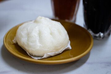 A freshly steamed Chinese pork bun served on a ceramic plate, with iced beverages in the background on a marble table.