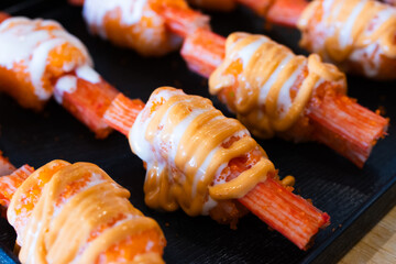 A imitation crab sticks topped with creamy mayonnaise and spicy sauce, arranged neatly on a black serving tray for Japanese-style appetizer or sushi.
