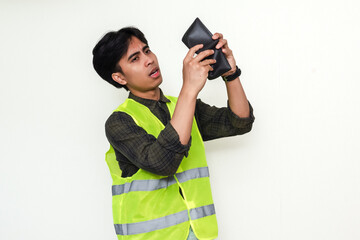 Fototapeta premium Sad Asian construction worker holding empty wallet, looking confused and hopeless. Isolated on white background. Concept of poverty, debt, job loss, and economic hardship